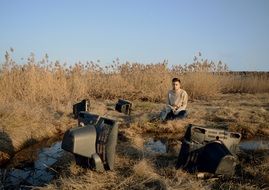 man sits among lapsed old televisions in wilderness