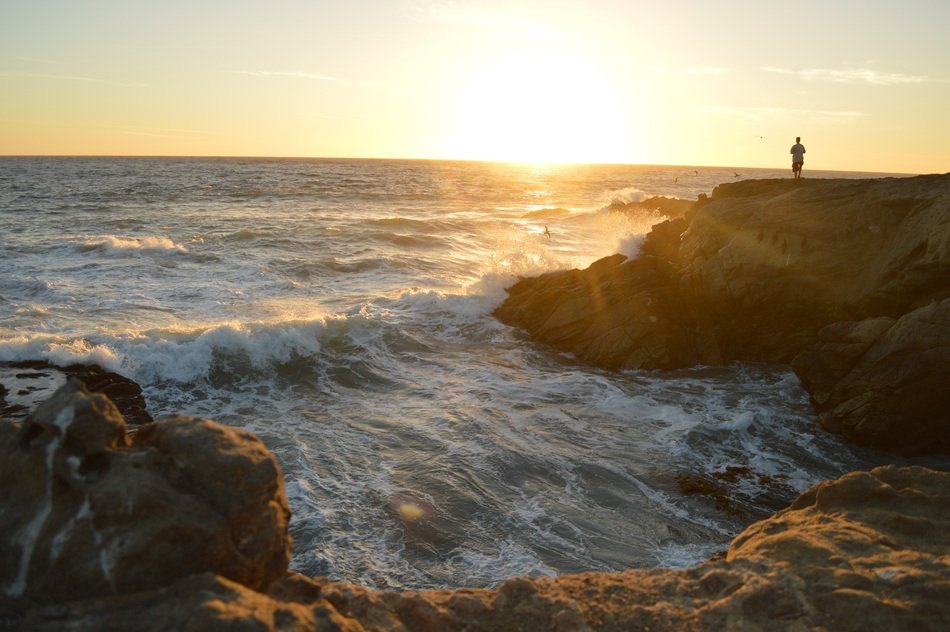 person on rocky Coast in front of foamy ocean at sunset