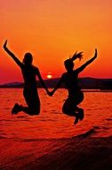 Silhouettes of the girls jumping near the water under the sunset