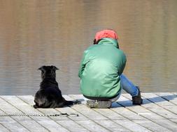 a woman in a green jacket and a black dog are sitting on a wooden floor on the shore of a pond