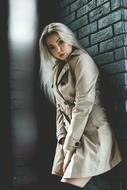 blonde in a light coat is standing against a gray brick wall
