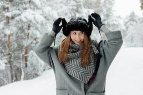 happy girl in a winter coat and a black hat on a background of snowy trees