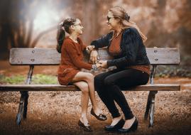 Smiling mother and daughter, with the glasses, sitting on the wooden bench, near the colorful and beautiful trees