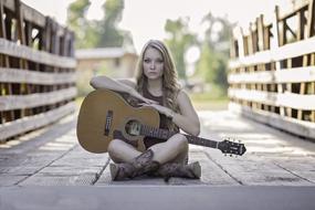 Portrait of a girl with guitar, sitting on the beautiful bridge in light, among the green plants, in summer