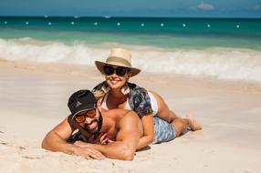 Smiling couple in sunglasses and hats, laying on the beautiful sandy beach, near the turquoise water