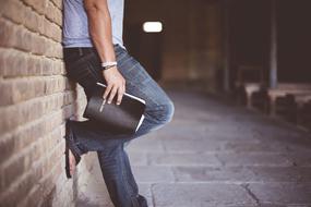 young man wearing Denim pants stands at brick wall with bible in hand