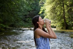 Profile portrait of the woman, drinking water from the glass, near the beautiful river, among the colorful trees