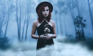 Gothic girl in black dress with white flowers in a foggy forest