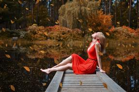 a girl in a red dress sits on a wooden pier on the shore of an autumn lake