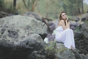 girl in a white dress sits on a boulder on the shore of the pond