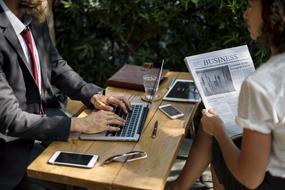 man typing on laptop computer and girl reading newspaper at table outdoor