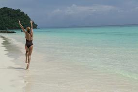 Back view of the girl in black swimwear, on the beautiful sandy beach, with the beautiful, turquoise water, on the island, near the green trees