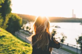 Back view of a blonde girl in sunlight, on the green hill, near the lake