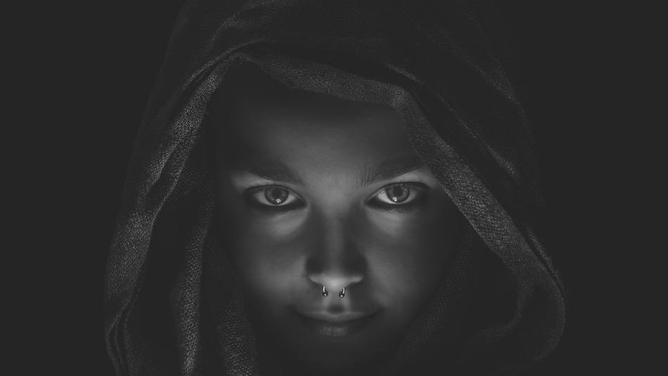 Black and white portrait of a woman, with the nose piercing, covered in the hood, among the darkness