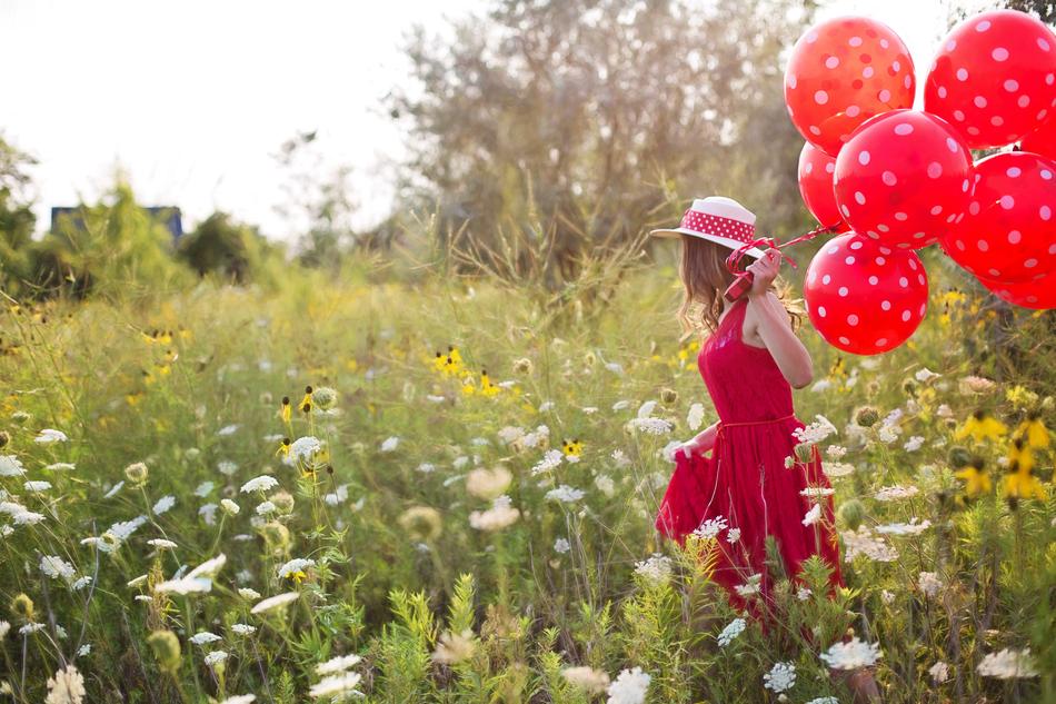 Young woman in dress and hat, with the red and white balloons, on the beautiful field with colorful flowers