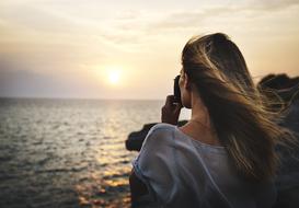 Back view of a blonde girl with the camera, on the beautiful coast, at colorful sunset on horizon