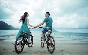 Back view of the couple, on the bicycles, on the sandy beach, near the beautiful mountains, under the cloudy sky