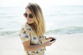 long haired girl in sunglasses stands with smartphone at sea
