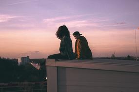 Girl and Guy sitting on edge of roof in city at dusk