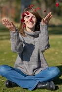 happy woman sits on lawn throwing up red petals in front of face