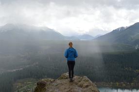 Back view of the woman in blue jacket, on the cliff, at the beautiful landscape with the mountains