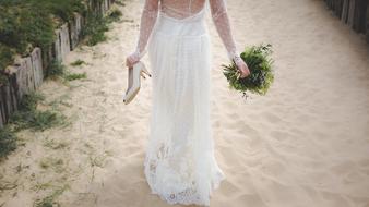 Girl in bridal dress walking barefoot on sand with shoes and bouquet