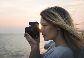 Profile portrait of the beautiful, blonde girl, taking a photo of the beautiful sea, at colorful sunset, with camera