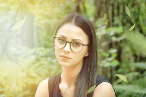 Portrait of the girl with long hair, wearing glasses, among the green plants in sunlight