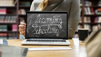 Person near the laptop with black board text "learning schooling, on the screen, in the library