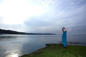 young woman in long blue dress posing on riverside with the hills at dusk