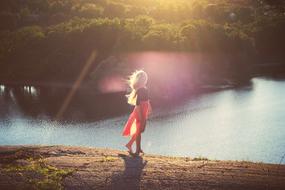 Blonde girl, standing on the rocky shore of the beautiful lake in sunlight, with green trees on the shore