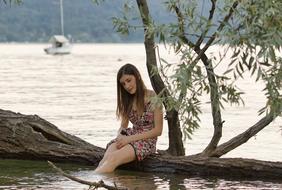 a girl sits on a tree trunk with her feet lowered into the lake