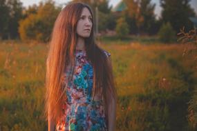 Red haired girl in colorful drees, posing among the beautiful and colorful field with trees in summer