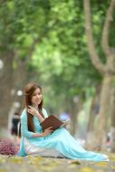 Beautiful Asian Girl in blue and white dress reading book among the trees in the park