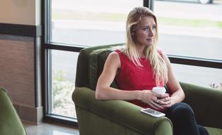 Blonde woman in red clothing, drinking coffee and sitting on the green armchair with the smartphone on it