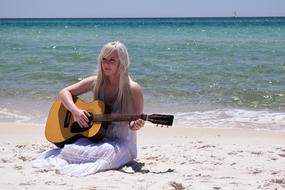 Blonde woman, playing on the wooden guitar, on the beautiful sandy beach with turquoise and blue water