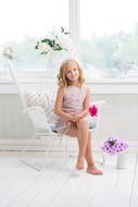 Smiling, blonde child girl, sitting on the white chair, with the colorful and beautiful flowers