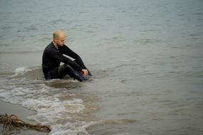 Male in black clothing, sitting in the water, on the sandy beach