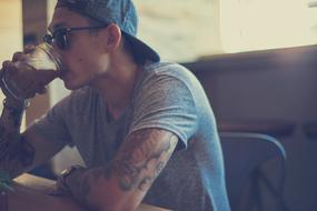 Guy with tattoos, wearing cap and sunglasses, drinking from the glass