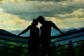 Black silhouette of a couple, near the fence, at beautiful and colorful background with mountains, under the cloudy sky