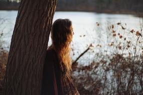 Girl posing near the tree on the shore with grass at colorful sunset