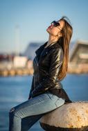 Posing woman in black jacket and sunglasses, posing on the beautiful shore with blue water