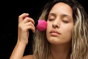 Portrait of a blonde woman, making make-up, using pink brush at black background