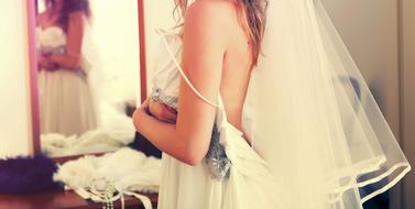 Back view of the bride woman in beautiful, white dress in front of the mirror