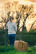 Back view of a blonde girl with the suitcase on the green meadow, near the trees, at colorful and beautiful sunset