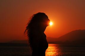 Silhouette of the curly girl, near the water, with the mountains, at beautiful and colorful sunset