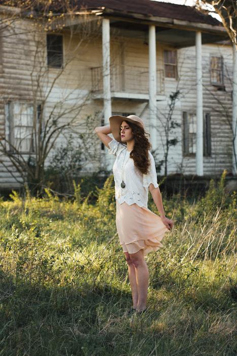 Posing woman in white and orange clothing, and in hat, on the beautiful green grass field, near the house