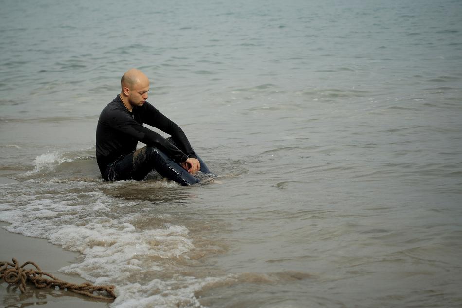 Male in black clothing, sitting in the water, on the sandy beach