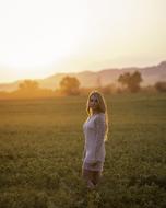 Blonde girl in white clothing, posing on the green field, near the trees and mountains, at colorful and beautiful sunset