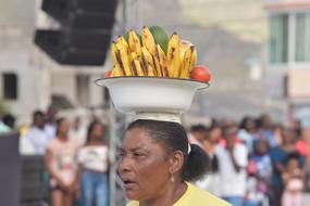 Black-skinned woman with the bowl with colorful fruits on the head, near the people